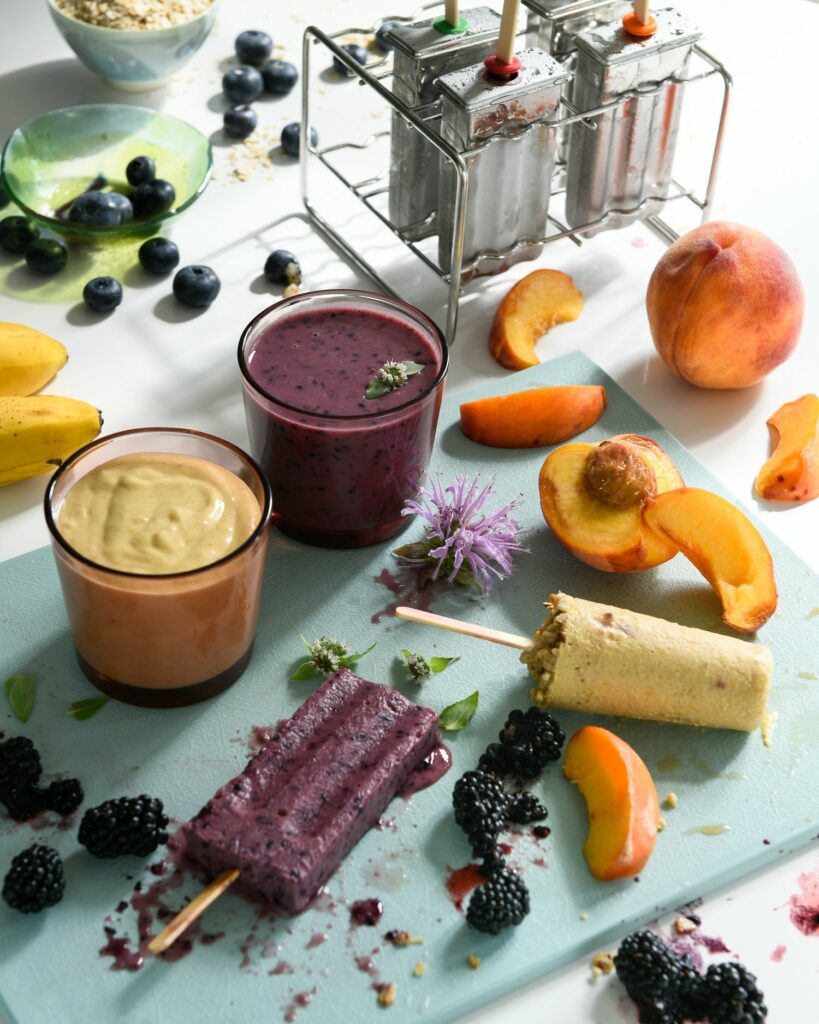A purple and green smoothie pop sit on a cutting board surrounded by fruit and two smoothies.