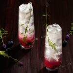 A lavender mocktail in a long, tall glass with maroon and lavender colors and a sprig or rosemary for garnish