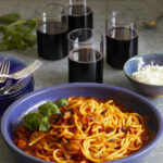 A Stanley Tucci Inspired Bucatini all' Amatriciana sits in a blue bowl with four glasses of wine placed behind the pasta.