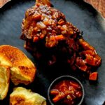 Beef short ribs covered in a boozy tomato sauce. The meat sits on a black plate with bread sitting to the left.