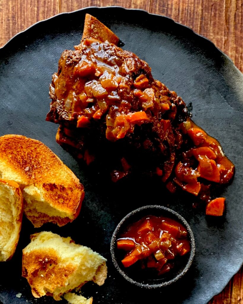 Beef short ribs covered in a boozy tomato sauce. The meat sits on a black plate with bread sitting to the left.