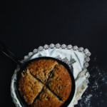 Buttermilk Soda Bread with Fennel Pollen in a frying pan placed on a napkin, which is placed on a dark black surface with sprinkled flour on it