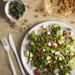 An aerial view of a Spring Time Salad with Saffron Vinaigrette, which sits on a white plate. A white fork and knife are positioned to the left of the salad.