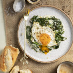 An aerial view of a dippy egg sitting on top of creamed spinach. a few pieces of sliced french bread sits off to the left of the plate.