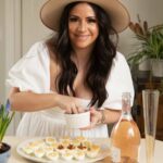 A woman in a white dress and wide brim hat smiles and leans forward as she sprinkles paprika over deviled eggs.