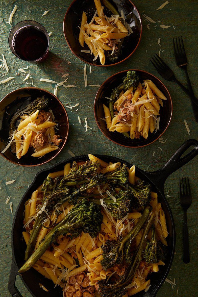 Penne with Broccolini and Chickpeas served in a frying pan and three bowls on a greenish surface with a glass full of dark liquid