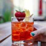 A Strawberry Negroni Bianco sits in a rocks glass, garnished with a strawberry and twist of a peel of lemon. A hand holds the glass on the right.