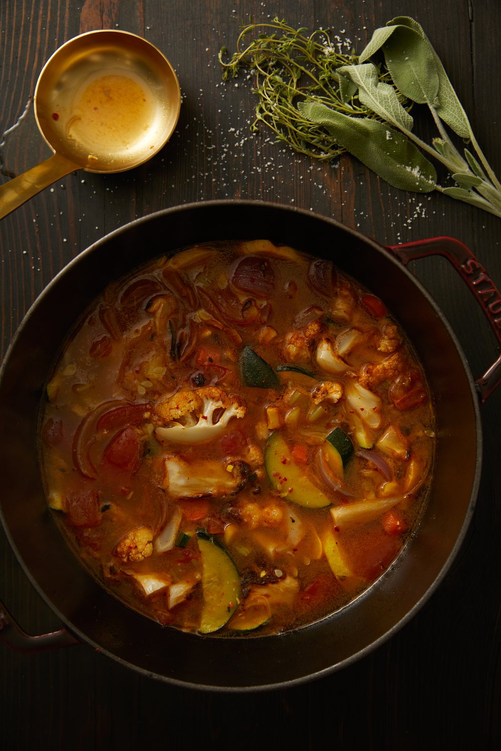 Vegetable Soup in a metal bowl with a spoon and green leaves on the sides, includes zucchini and yellow squash.