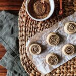 An aerial view of five cinnamon rolls on a piece of parchment paper next to a bowl of cinnamon with a spoon in it.