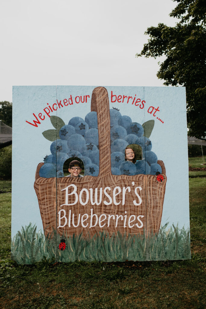 A painted sign with a blueberry basket and text that says "We picked our berries at Bower's Blueberries." Two blueberries have spots for faces, and two kids stick their heads through. 