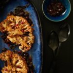 Cauliflower Steaks with Curry Leaf Oil and Red Wine Cherry Sauce sit on a blue plate to the left of the frame.