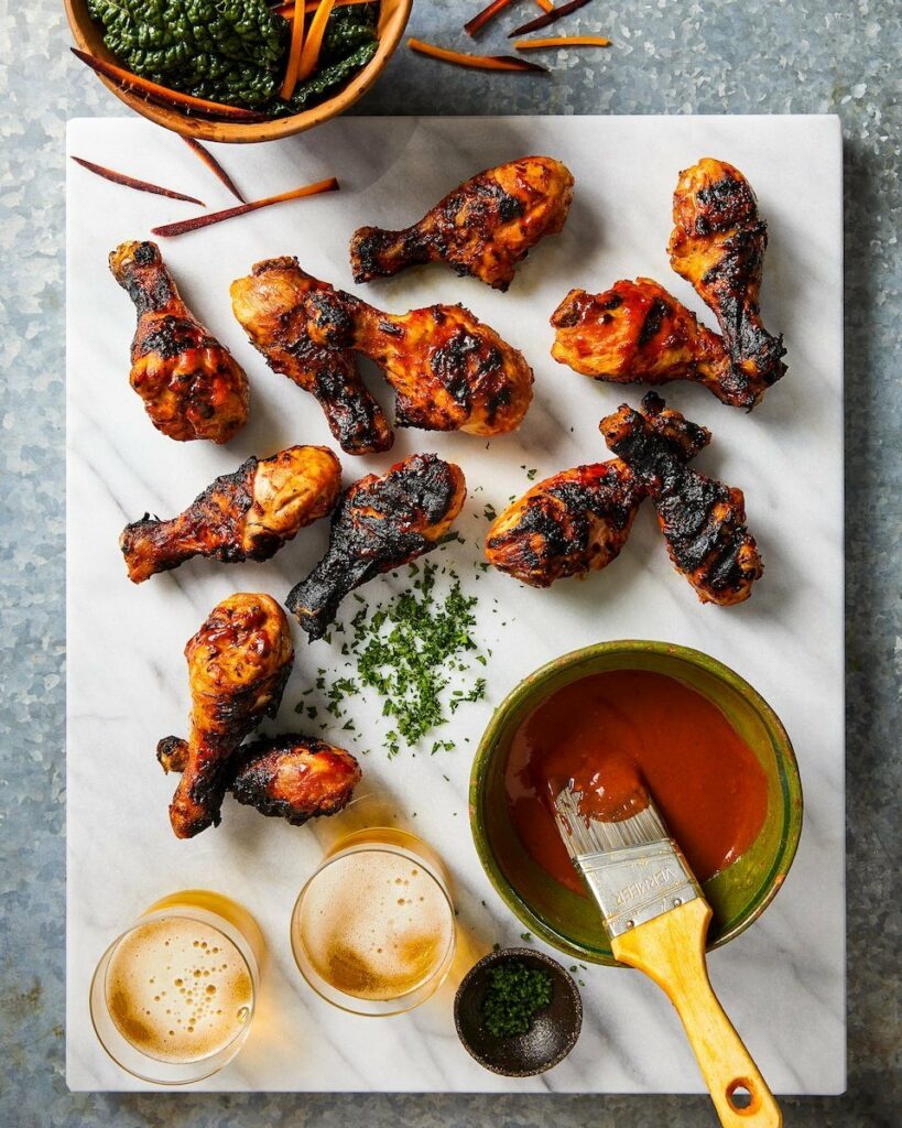 10 grilled wings with even charring sit on a white marble cutting board alongside two beers and a bowl of homemade bbq sauce.