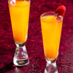 2 tall narrow orange cocktails with a pick sugar petal and raspberry garnish with a deep red background, Taylor Swift cocktails