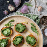 green nasturtium pesto on crostini with small cucumber slices on a beige tray garnished with arugula, a black background, green floral fabric, tan burlap and fresh garlic with purple skins