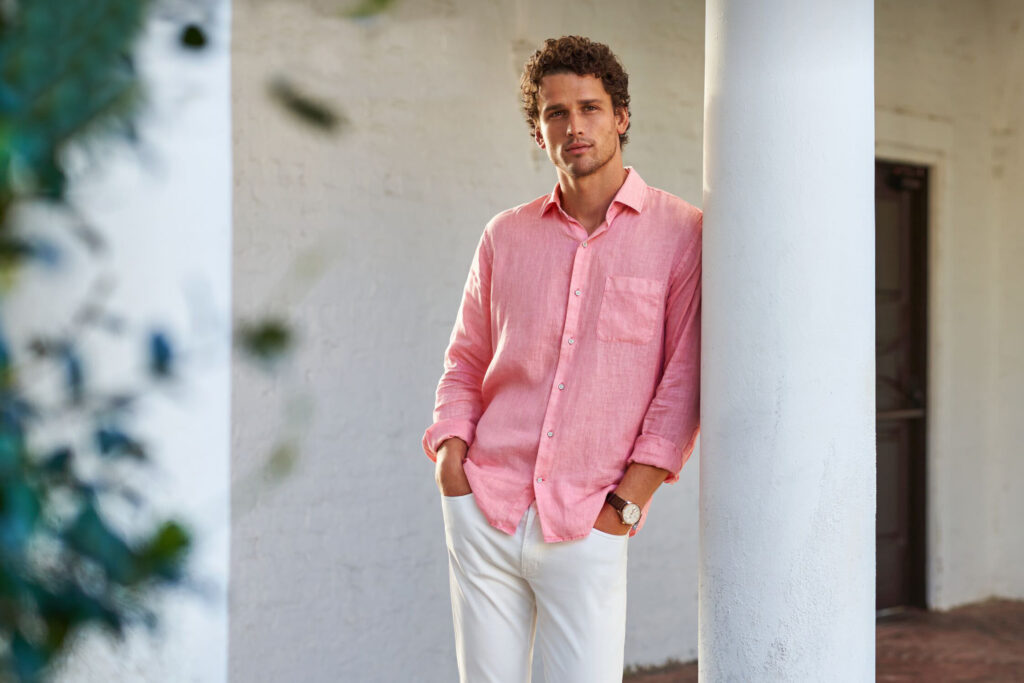 A white man leans against a white column in a pink linen shirt and white pants