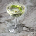 a lemon basil martini cocktail in a coupe glass sitting on a white and grey marble slab with fresh basil and olive oil floating on the top of the cocktail