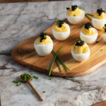 a wooden board on a piece of white and gray marble on a wooden table with six deviled eggs topped with caviar and garnished with chives