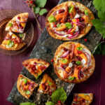 An aerial view of three Peach Wineberry Flatbreads, with one cut into six slices. They are bright in color and sit on a black wooden slab. Peach Wineberry Flatbread Recipe