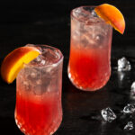 a look in at 2 identical dark peachy pink cocktail on a black background with ice cube and peach wedges as garnish