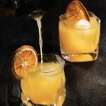 2 orange cocktail in rocks glasses on a black surface with dehydrated orange slices as garnish. The front cocktail is being poured by a woman's hand