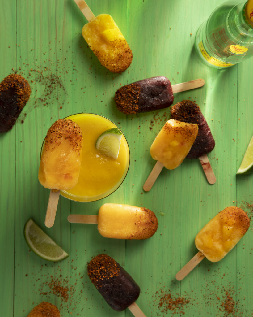 Various dark red and orange colored fruit popsicles sit on green wood. Paleta Popsicles Recipe