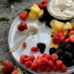 Berries and pineapple on a white plate with a bowl of creamy white fruit dip, and a little gold spoon with some of the dip on a plate