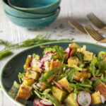 Three individual blue bowls above a green serving bowl filled with a vibrant potato salad of yellow, green, red, and purple ingredient