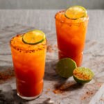 Two Michelada cocktails orange red in color sit in tall glasses, garnished with lime and Tajín. Michelada recipe