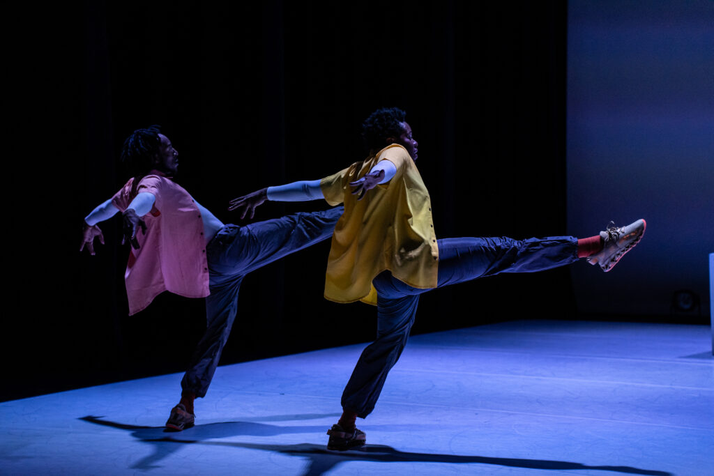 two dancers in loose fitting clothing hold their right leg up with their arms facing back