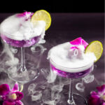 Two glasses with purple liquid inside are topped with little botanical pink flowers and lemon slices with smoke pouring over the cocktail.