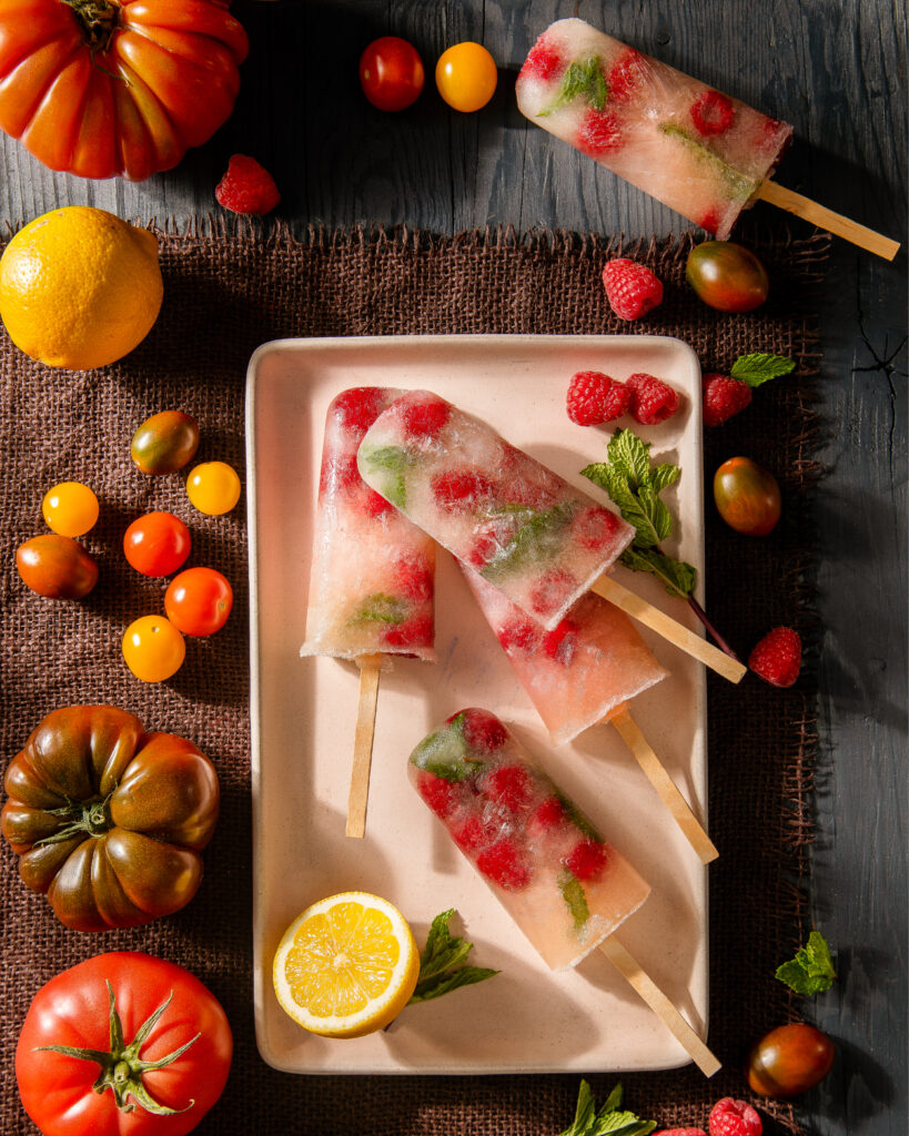 Four Tomato Water Lemonade Popsicles with Raspberries & Mint sit on a white tray surrounded by tomatoes of various colors and sizes