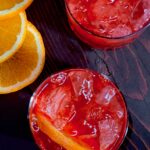 A close down shot of two red cocktails garnished with orange and a fan of orange slices at the top left.