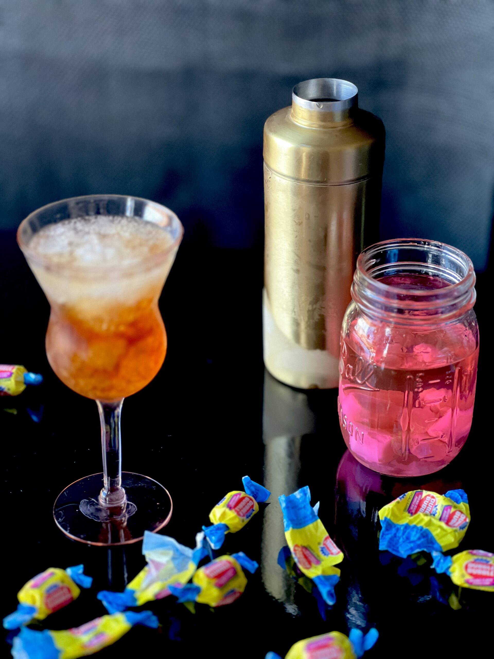 A stemmed glass with a light cola colored cocktail, a gold cocktail shaker and a mason jar with a bubble gum pink vodka, surrounded by bubble gum and bubble gum wrappers