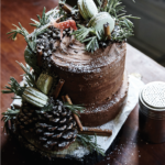 A Whole Grain Apple Cider Layer Cake covered in chocolate frosting and white decor like pinecone and cinnamon sticks.