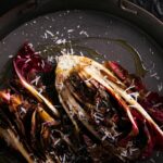 A head of radicchio cut in half and charred on a dark plate with shaved parmesan.