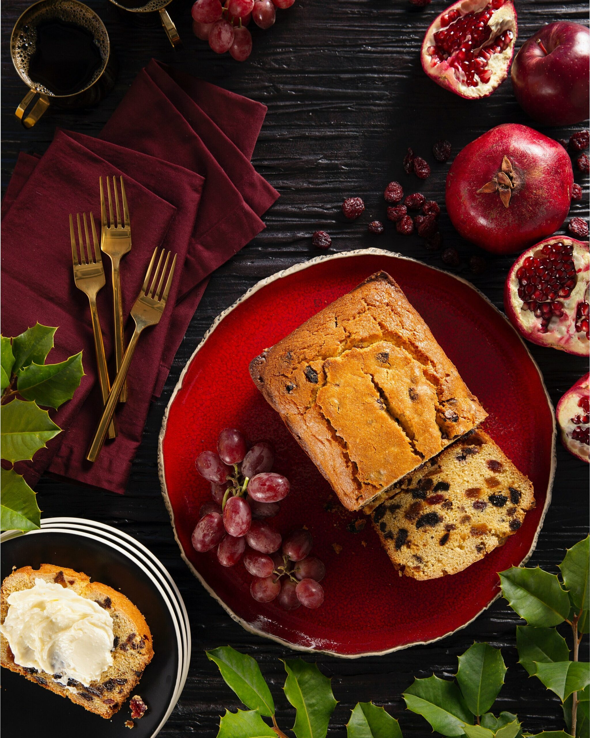 An aerial shot of a Polenta Fruitcake Quick Bread on a red plate, surrounded by raisins, grapes, and pomegranates. Polenta Fruitcake Quick Bread Recipe