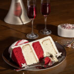 A slice of peppermint cake, white and red in color, sits on a plate next to two champagne glasses of red liquid.