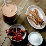 A clear glass mug of spiked hot chocolate with a small plate of dried red ancho chilis, a small oval dish of cinnamon sticks, and a small bowl of salt on a wooden surface