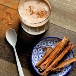 A creamy brown beverage with a sprinkle of cinnamon sits in a clear glass. cinnamon sticks on a plate with blue floral pattern sits to the right. Keto Pumpkin Spice Frosty