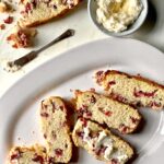 An aerial view of a lemon keto cranberry quick bread sitting chopped up into five slices on a white plate. the middle slice has butter on it. A small bowl of whipped butter sits to the top right.