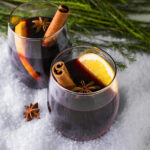 Two stemless wine glasses with mulled wine sitting on a snowy surface with pine greens, garnished with a cinnamon stick, star anise, and an orange slice.