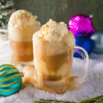 Two boozy root beer floats I frosted mugs overflowing with frothy root beer on a snowy surface with colorful Christmas ornaments and greenery.