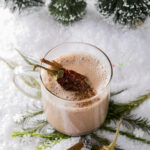 A clear mug of hot chocolate on a snowy surface with evergreen clippings and dried ancho chili peppers.