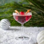 A cocktail glass filled with Rudolph's Red-Nose Punch topped with a cranberry and pretzels on the glass.