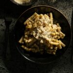 A dish of creamy pesto sauce pasta in a black bowl beside parmesan cheese made by Victoria Sande.