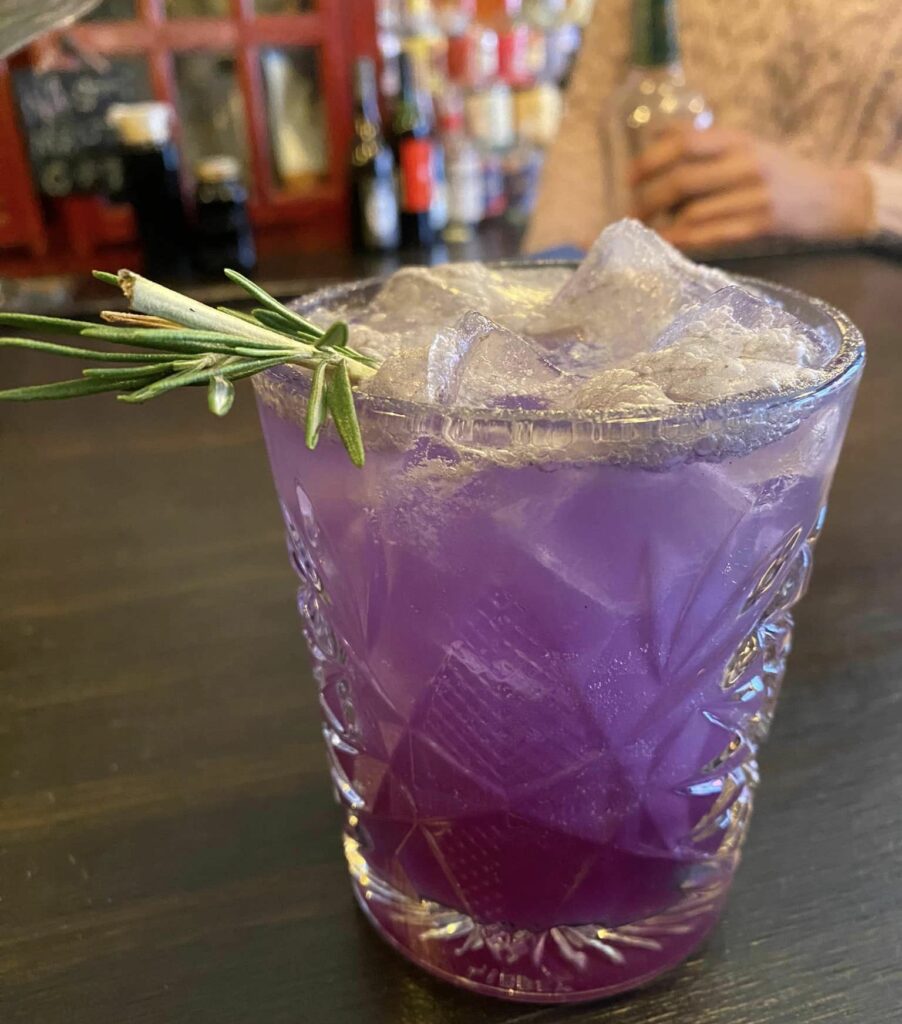 a photo of a non alcoholic purple cocktail with a sprig of rosemary.