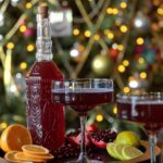 A fancy bottle with a deep red Good Luck Sparkling Pomegranate Cosmo on a tray with 2 cocktail glasses, orange slices, an open pomegranate, and lemon slices in front of a blurred Christmas tree.