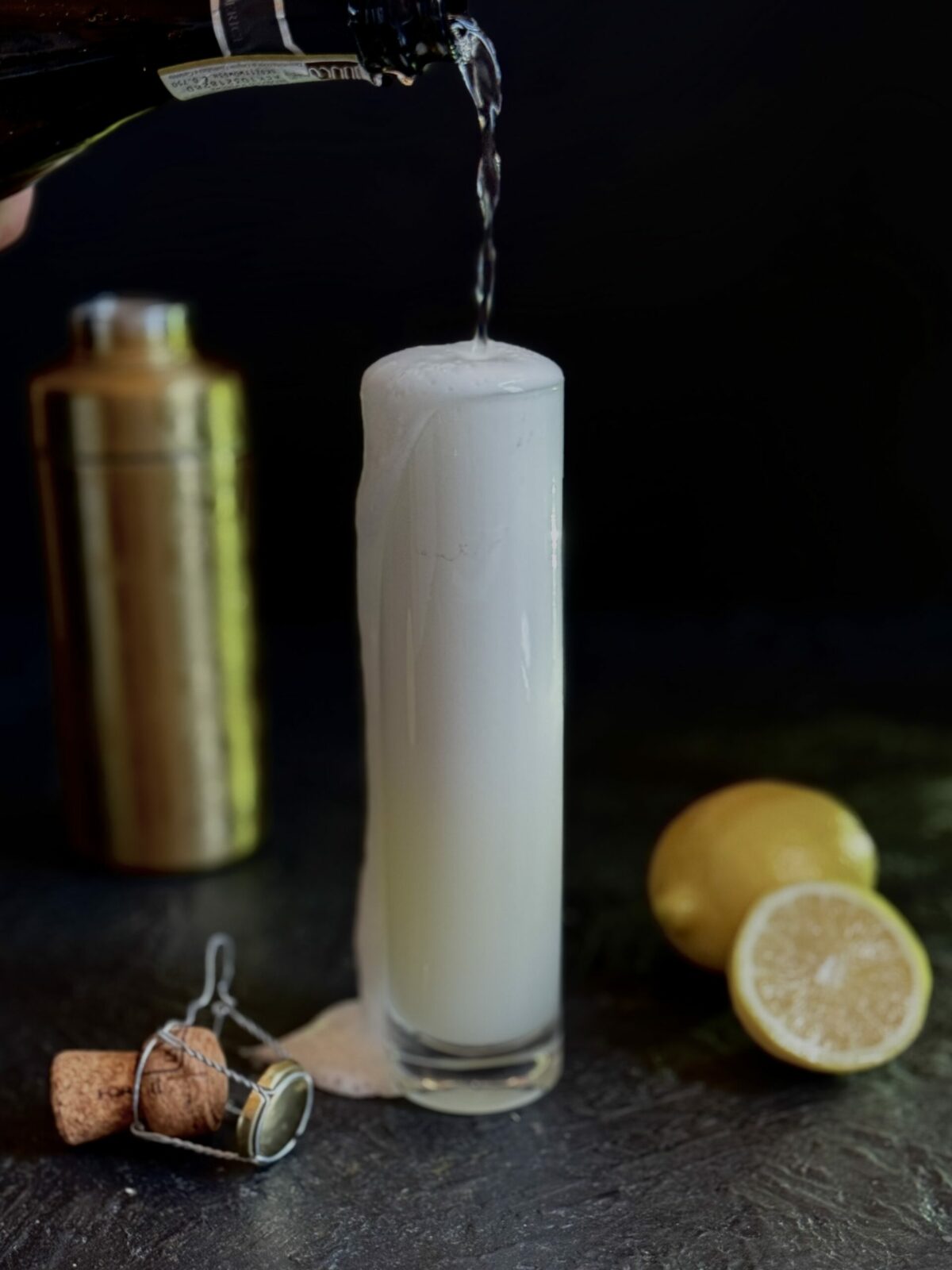 The Gin Fizz with Rizz: A tall, thin highball glass with a cocktail topped with champagne on a black background with a gold cocktail shaker, lemon, and the bottle cork as styling elements.