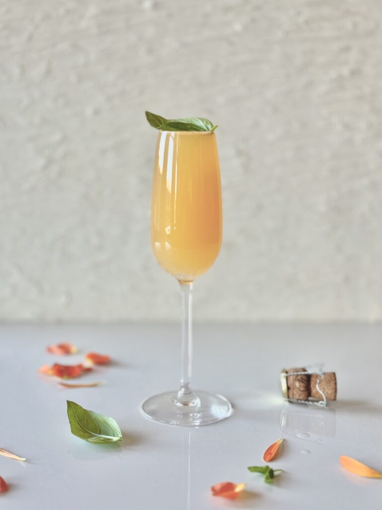 A champagne flute with a peach cocktail garnished with a green basil leaf on a white surface with a champagne cork, basil leaves, and peach and orange flower petals for styling.