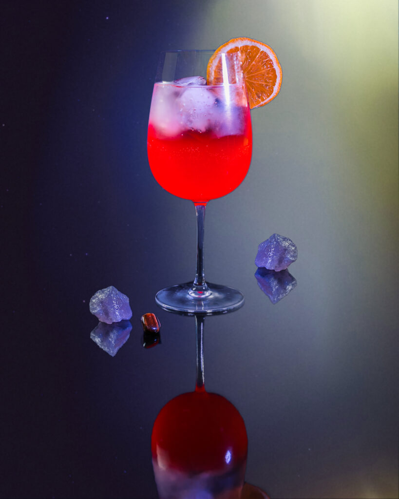 A cocktail for Leo with red liquid in a wine glass topped with an orange slice and surrounded by crystals on the table.